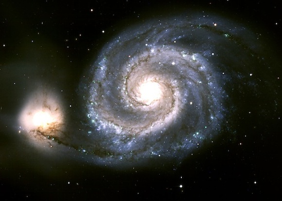 Spiral galaxy shown as bright central light and surrounding spiral arms, with a companion irregular (bright light) galaxy on the left side.