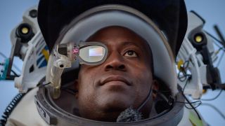 A man wears a eye covering over his right eye and stares off into the distance. It looks like they are wearing some sort of spacesuit.