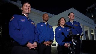 four humans in blue jackets smile before microphones, in front of a white building.