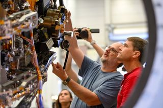 two people peering at an open spacecraft door that has a lot of gears and mechanics on it