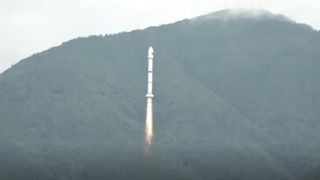 a white rocket launches into a cloudy sky with a forested mountain in the background