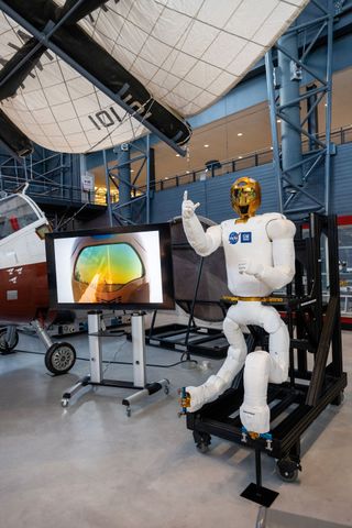 a white humanoid robot with a gold helmet sits in a museum exhibit hall