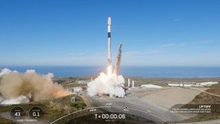 a black and white rocket launches into a clear blue sky from a seaside pad