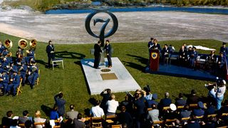 a crowd of people watch as two military men shake hands while standing beside a monument that features the number 7.