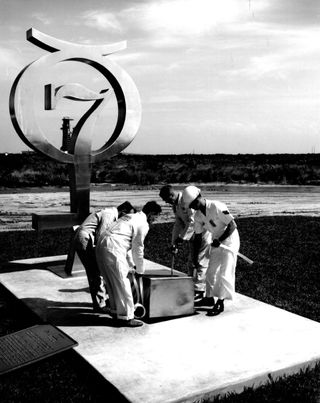 black and white photo of four people lowering a metal box into the ground next to a monument featuring the number 7