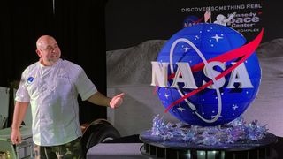 a man in a white t-shirt stands next to a large spherical cake that looks like nasa's famous