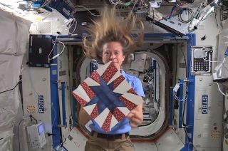 a woman with blond hair holds up a red, white and blue quilt on board the international space station