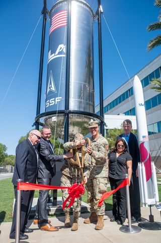 two people in military fatigues use oversize novelty scissors to cut a red ribbon in front of a cylindrical blue rocket propellant tank outside an office building