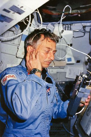 a man in a light blue flight set listens to a radio in a cramped room filed with wires and computers