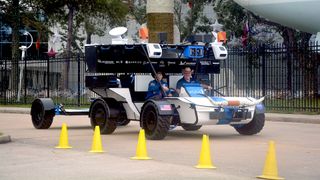 two people drive a large, six-wheeled moon rover on a sidewalk bordered by yellow cones