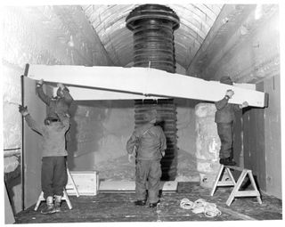 Four men, two on the left, one in middle, one on the right hoist a cross bar support beam in a tunnel. Behind them, a tall ribbed black pipe stands from floor to ceiling.