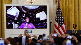 People's hands are raised holding phones in the foreground, to record a large screen next to an American flag, in front of a gold curtain. on the screen, a woman in a pink shirt holds a microphone below a window on a space station.