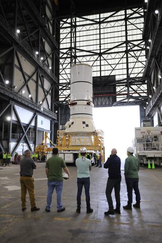 a rocket piece on top of a cart inside a large warehouse. several people look at it with their backs to the camera