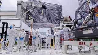 scientists in white clean suits move a metallic honeycomb-like structure onto a rectangular piece of electronics covered in wires and components