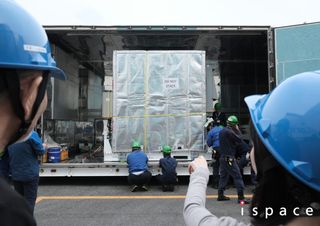 the back of the heads of two blue-helmet wearing people are in the foreground, with one pointing at a large metal box being lifted off a truck by two kneeling people in green helmets.