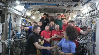 A man in a black shirt shakes the hand of a woman in a blue shirt, inside a crowded room of wires and electronic gear. behind them, six people, four in red, one in grey, one in gree, one in black, and the last in blue.