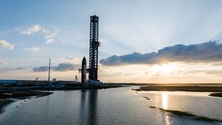 a large silver spacecraft sits next to a launch tower near the sea, with the rising sun in the background