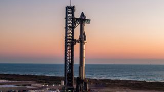 a giant silver rocket stands vertically at its seaside pad near dawn