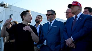 a man in a black t-shirt talks with several men wearing suits.