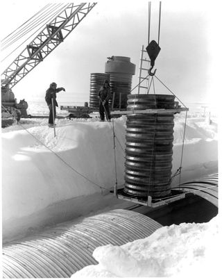 a crane on the left lowers a ribbed pipe on the right, attached with a large hook, lowing into an open section of embedded structure in the snow.