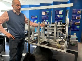 A man in a salmon shirt and blue sweater vest places in left hand on a metal framework with two fingers of his right hand on his hip.