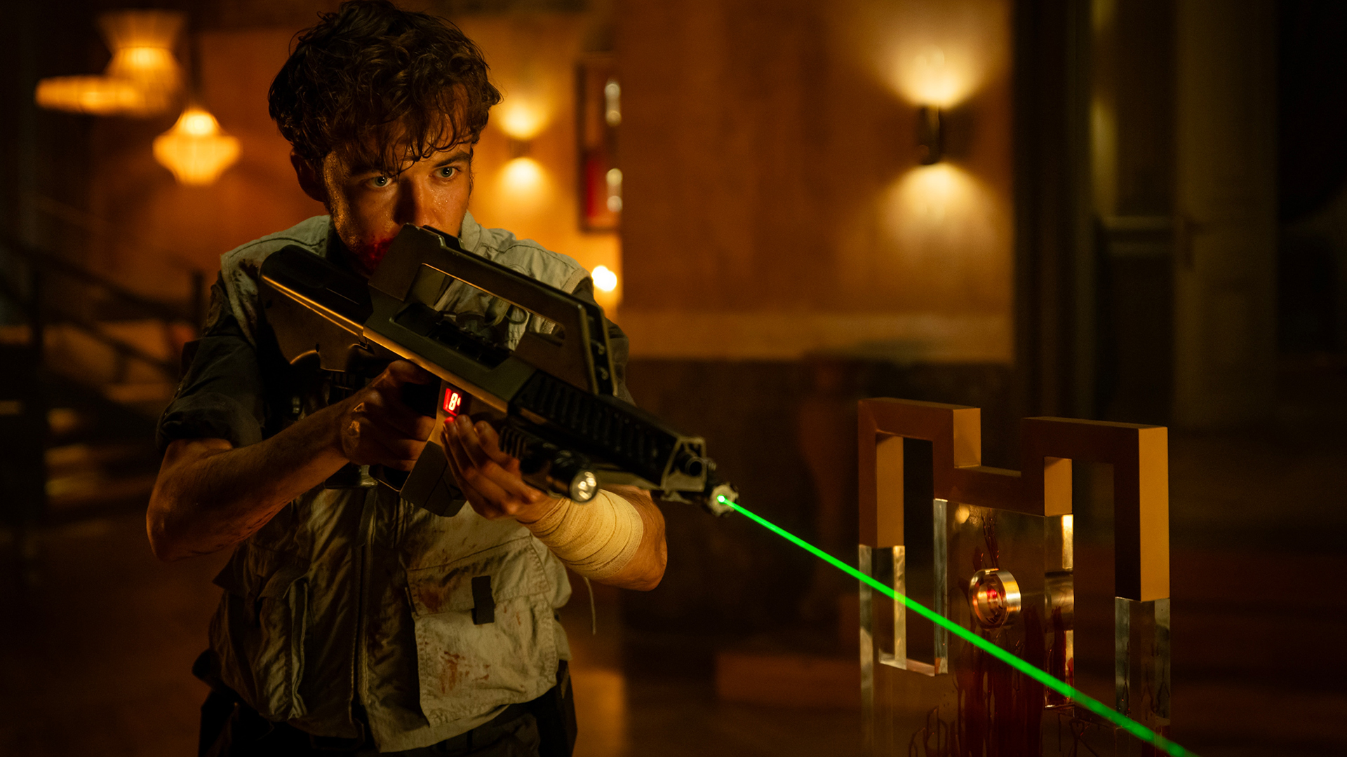 Screenshot from Alien: Earth. A young man holding a sci-fi assault rifle with a green laser sight affixed to the front. He's in an ornate room, with blood splattered around.