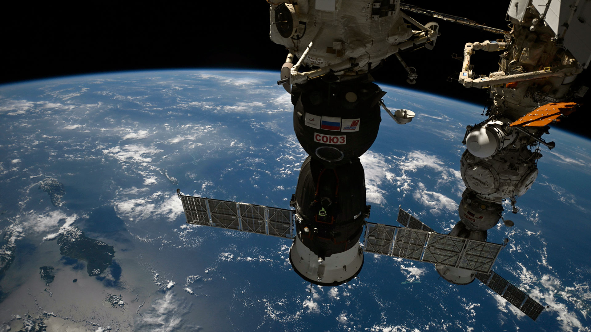 view of two space capsule docked side by side at a space station over a blue and white planet Earth