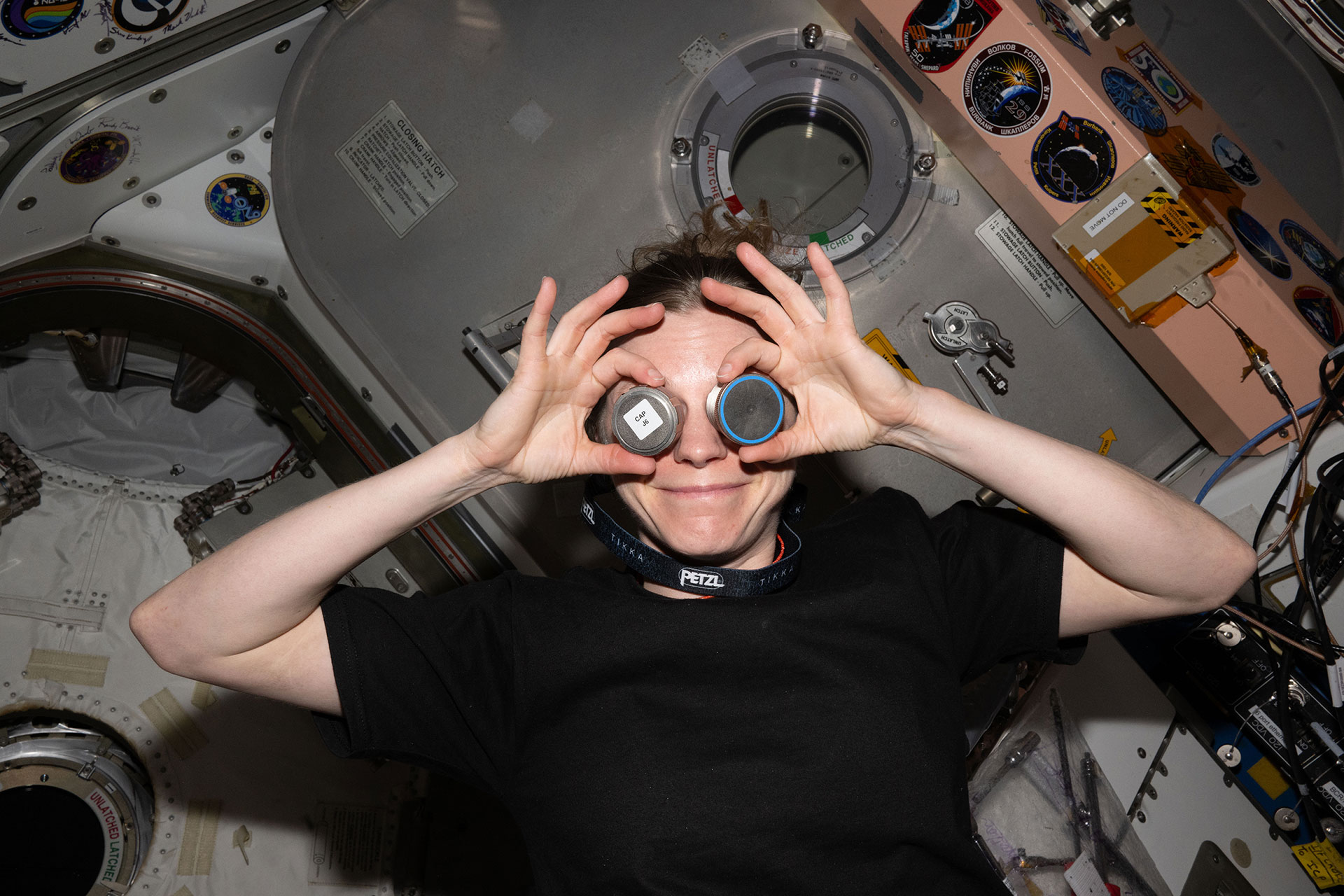 a woman wearing a black short sleeve shirt playfully holds up hardware covers to her eyes as if they were a pair of goggles on board a space station