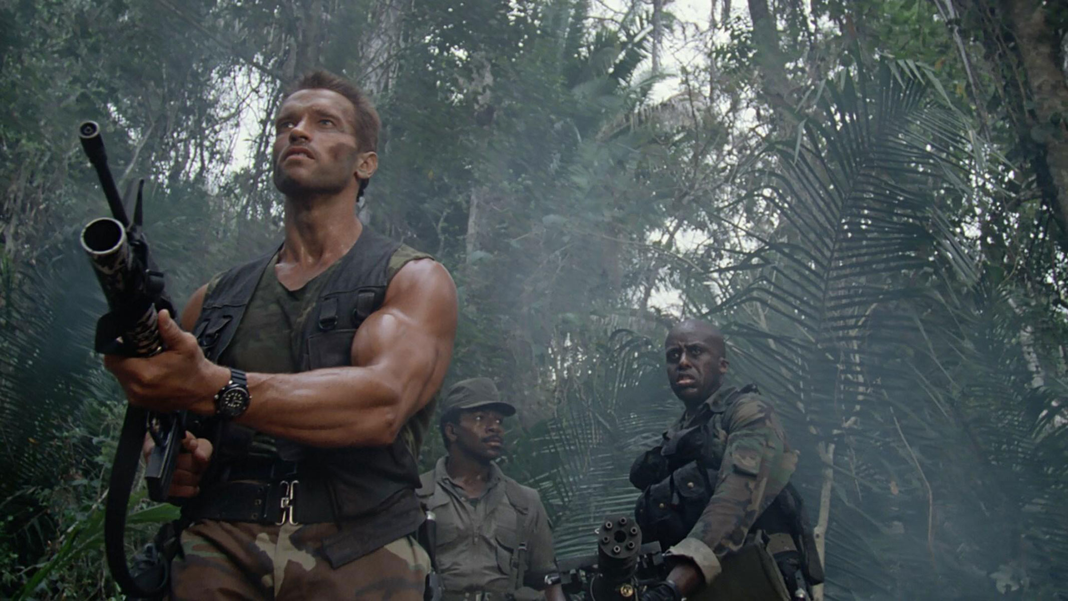 A man with large muscles is holding a gun, while a couple of other men in military gear stand behind him, all among some trees.