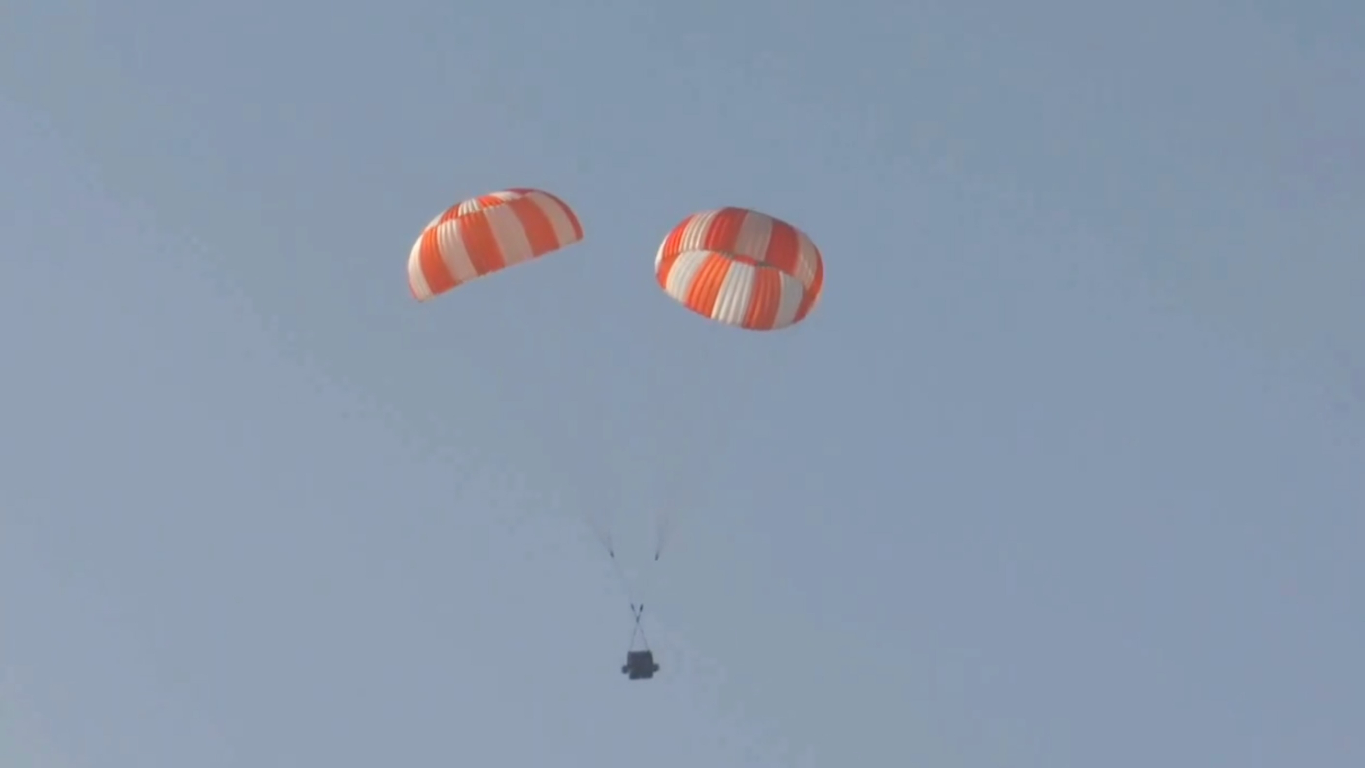 a mock space capsule descends through blue skies under two orange and white parachutes
