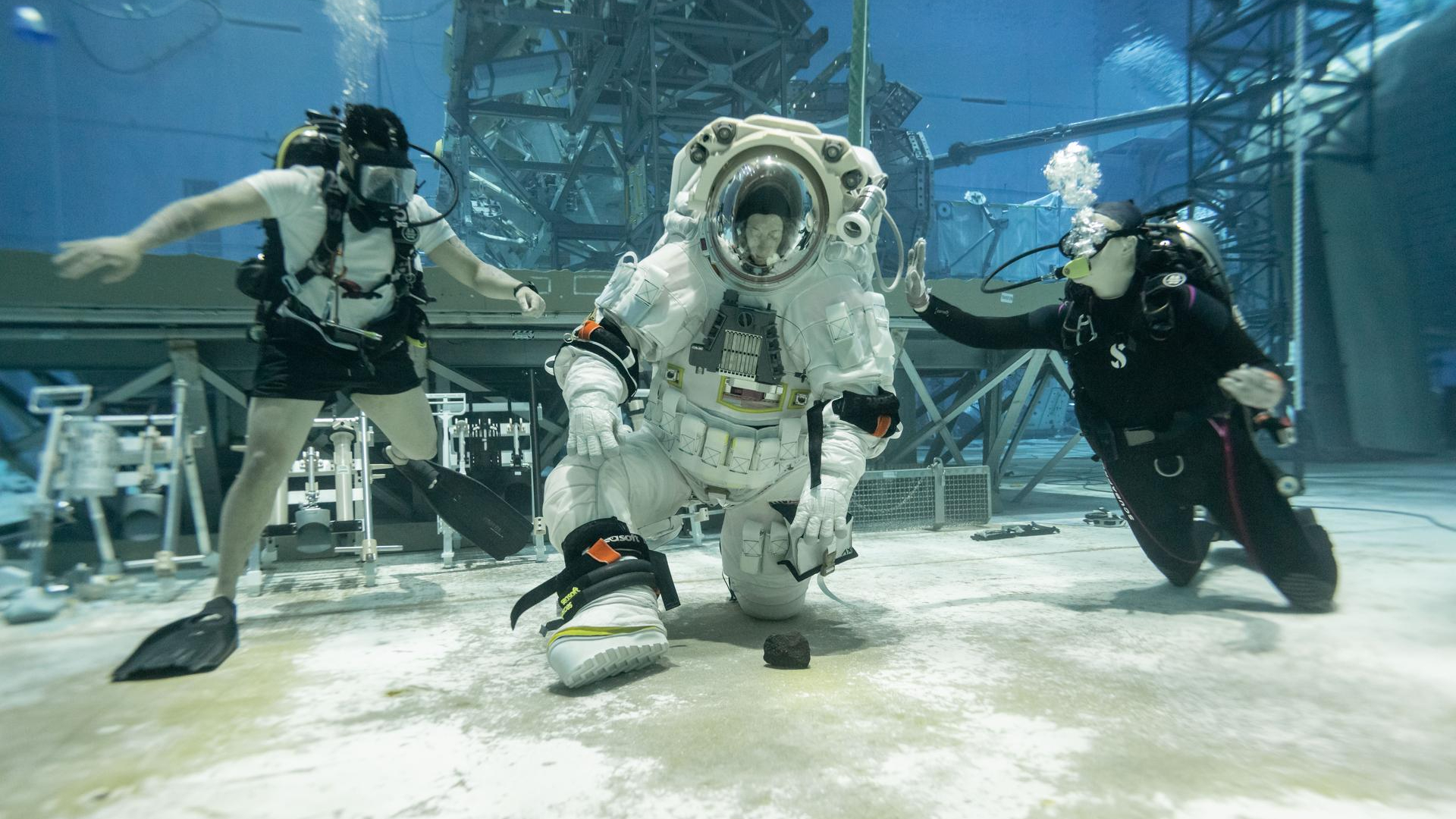 A person wearing a white spacesuit with a clear helmet kneels down to pick up a rock at the bottom of a large sandy pool with two people wearing dark swimsuits on either side of them