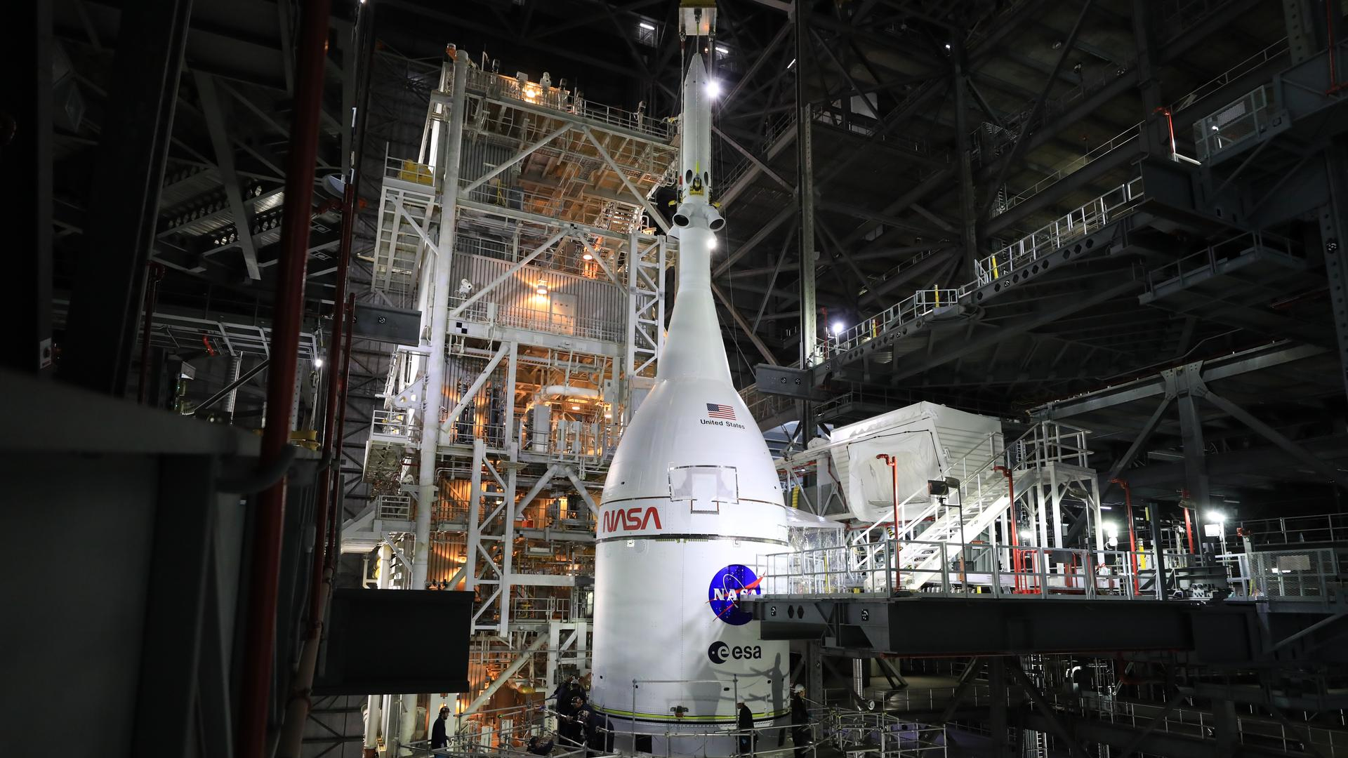 A white pointed rocket module sits on top of a white cylinder, both having the red worm NASA logo and the blue and red meatball NASA logo. The entire system sits in a large warehouse