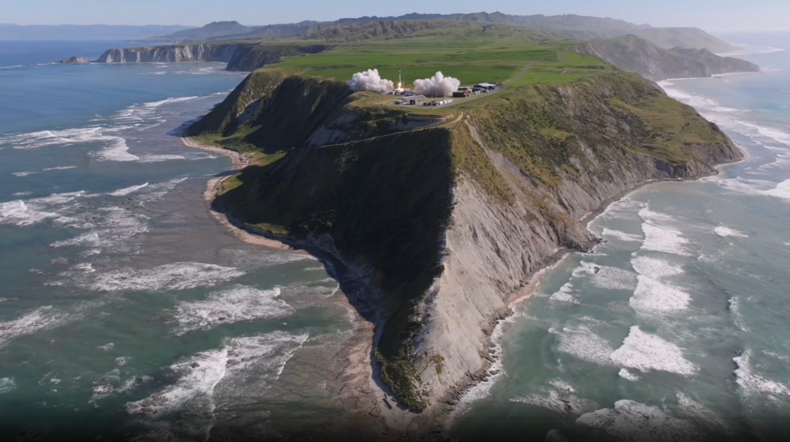 a plume of grey smoke forms on the tip of a rocky island rising out of a wavy ocean