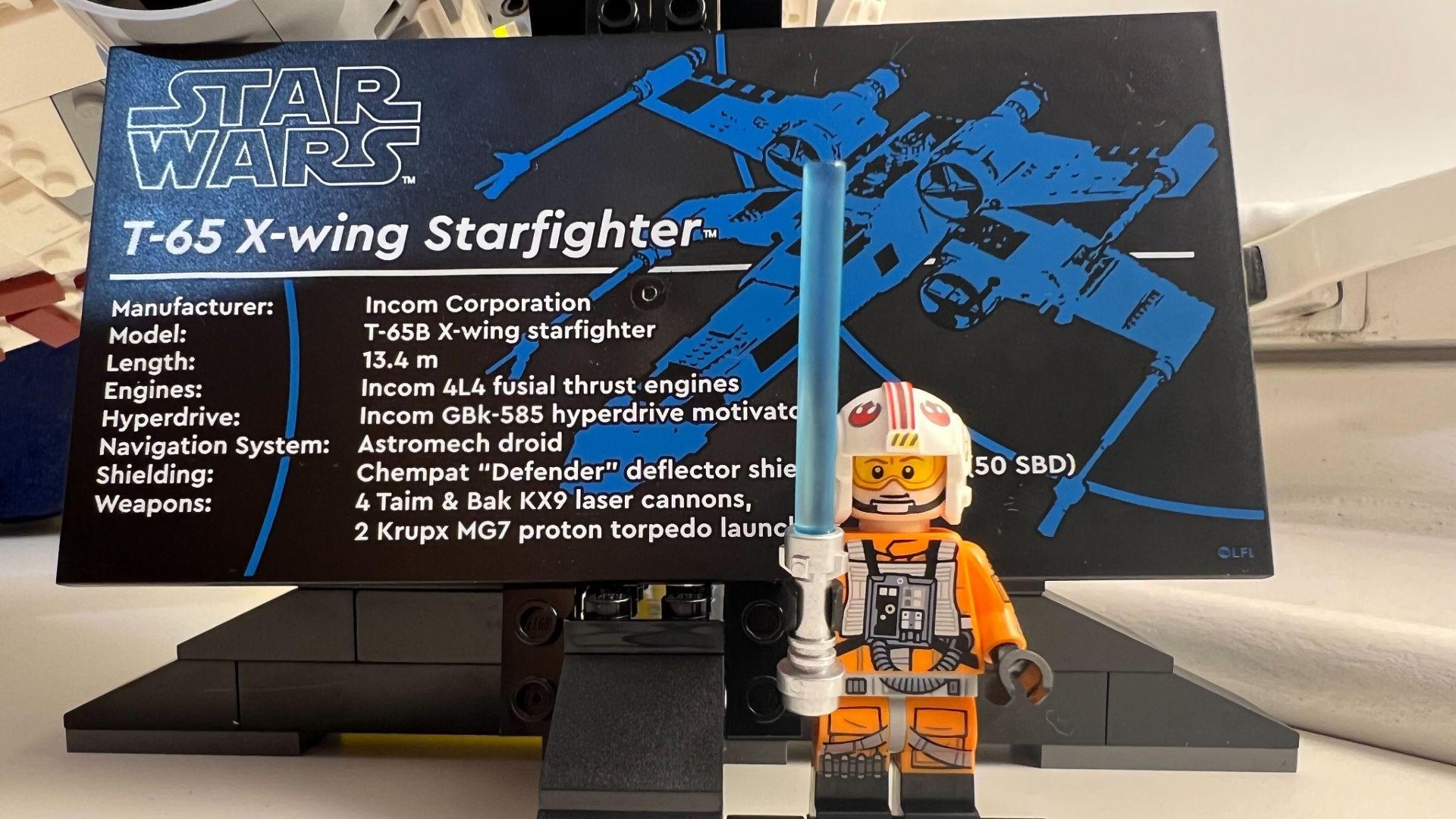 Lego Star Wars X-Wing Starfighter 75355-plaque front view with Rebel Pilot Luke minifigure standing in front holding blue lightsaber.