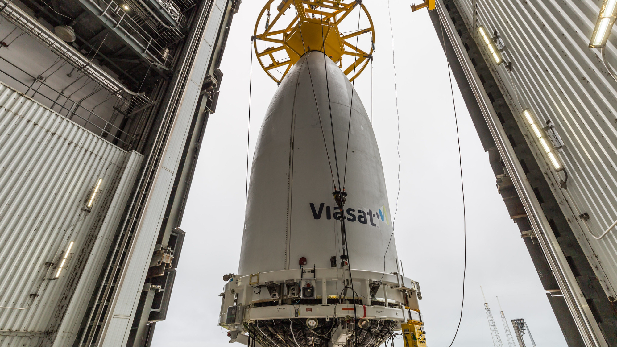a white conical payload fairing is hoisted by a crane inside a hangar