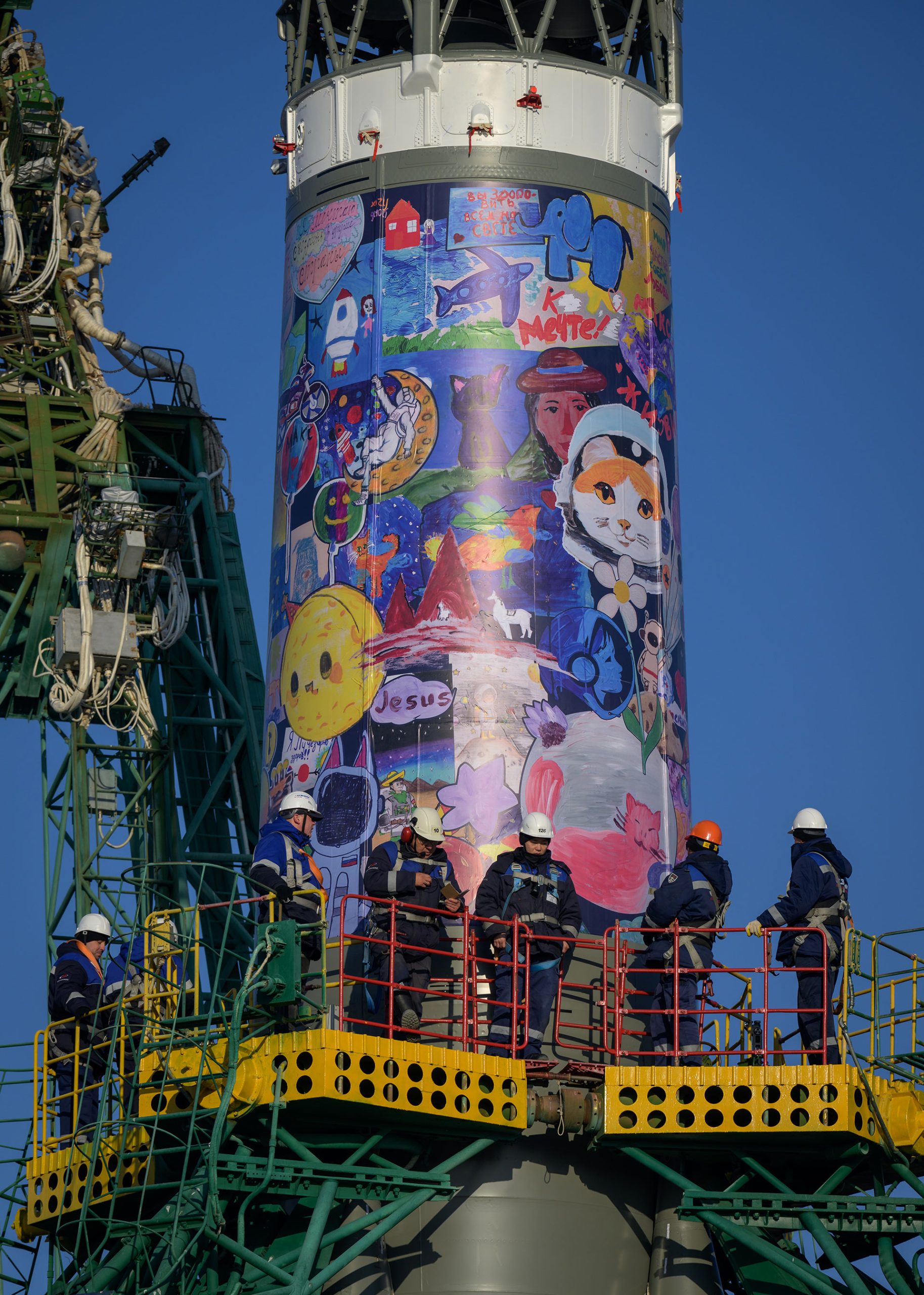 a colorful mural featuring children's drawings is seen wrapped around the body of a rocket on its launch pad