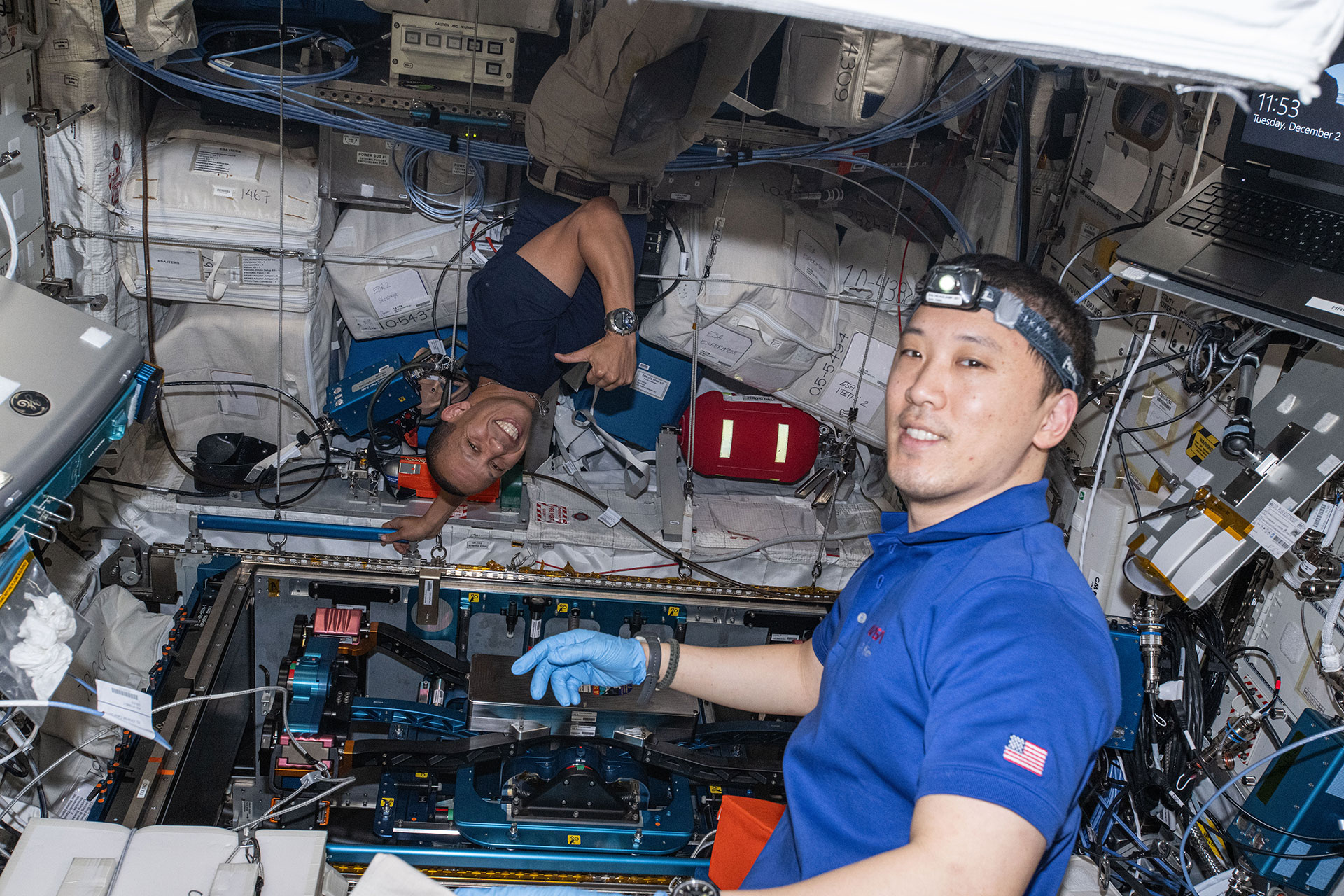 two men, both in blue short sleeve shirts and one wearing silicone gloves and a head lamp, work together to install equipment aboard a space station