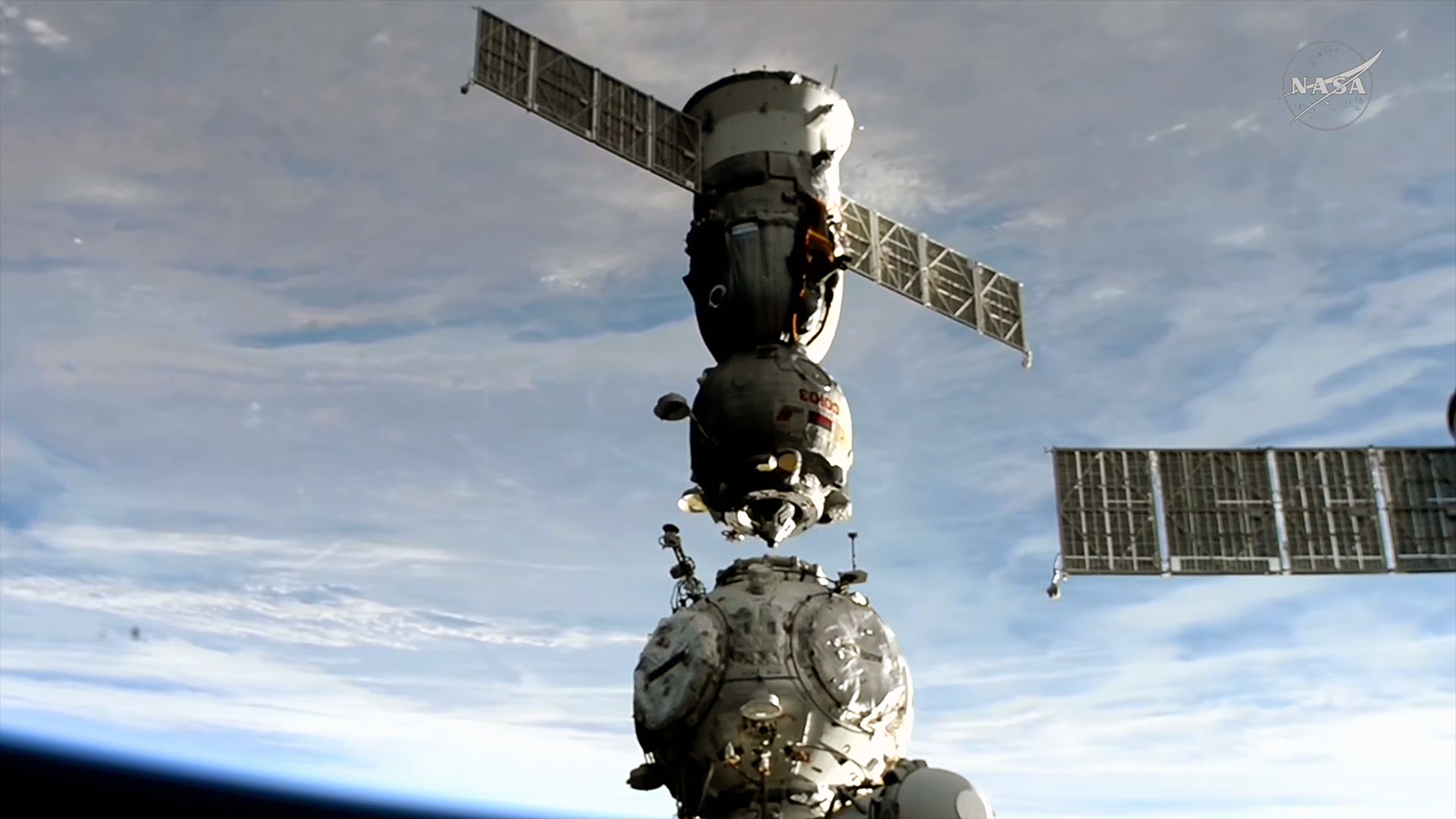 a spacecraft separates from its docking port at a space station backdropped by the blue and white of the Earth below.