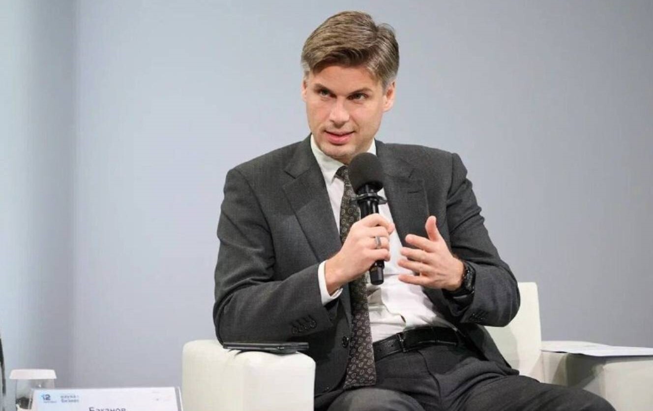 A man with gray hair wearing a black suit and slacks and a white button up shirt sits in a white chair and holds a microphone