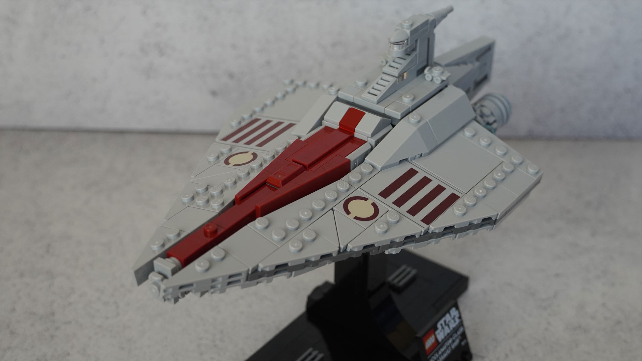 A miniature-sized long grey ship made of bricks with a red stripe running down the middle is placed on a black stand in front of a grey background.