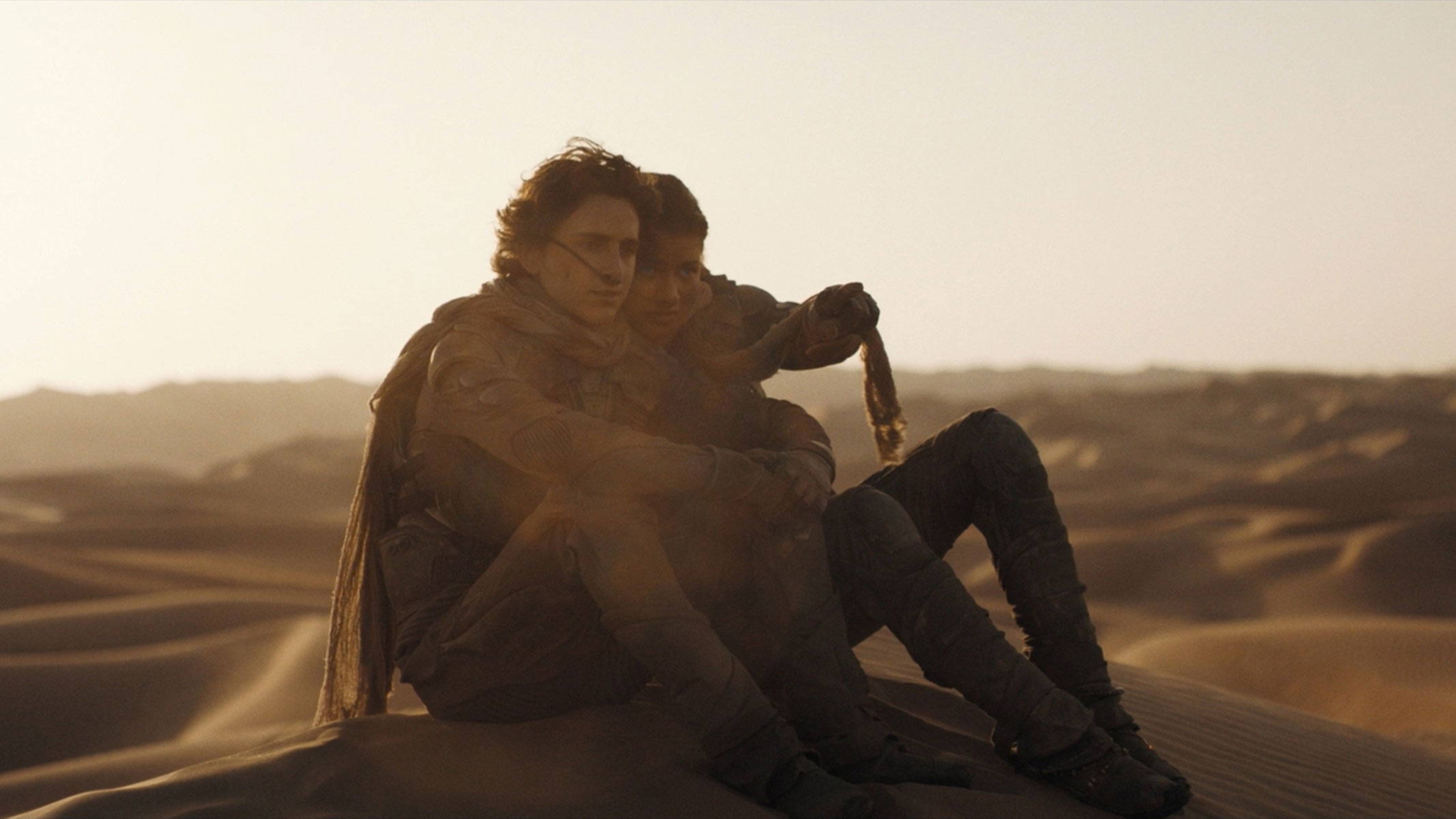 A man and a woman sit on top of a sand dune with a lowering sun in the background.