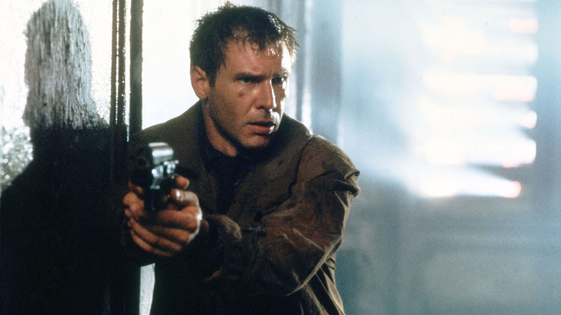 A man is holding a gun next to a concrete wall with a metallic background behind him.