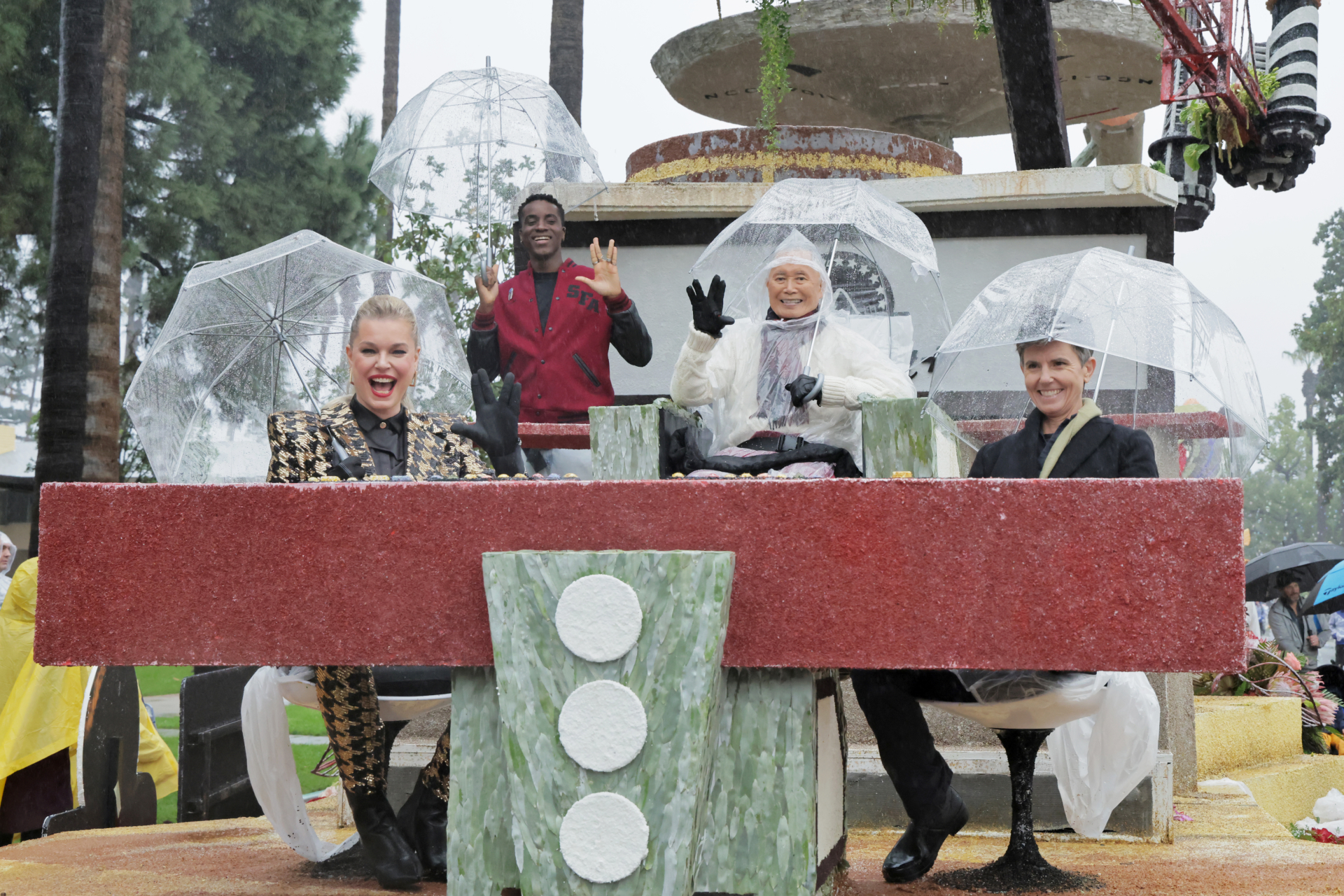 Four people sit on top of a parade float with clear plastic umbrellas. They all wave at the camera.