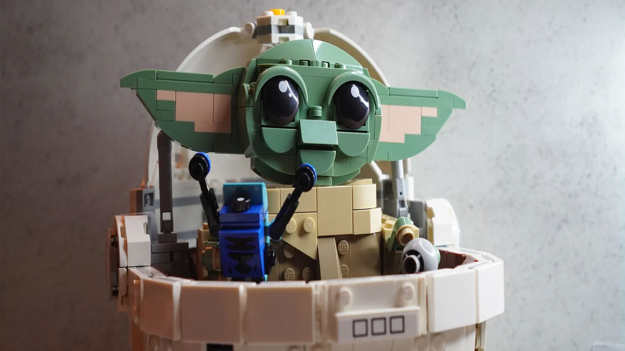 A green creature in Lego form with giant ears is sitting in a white pram, also in lego form, infront of a grey background