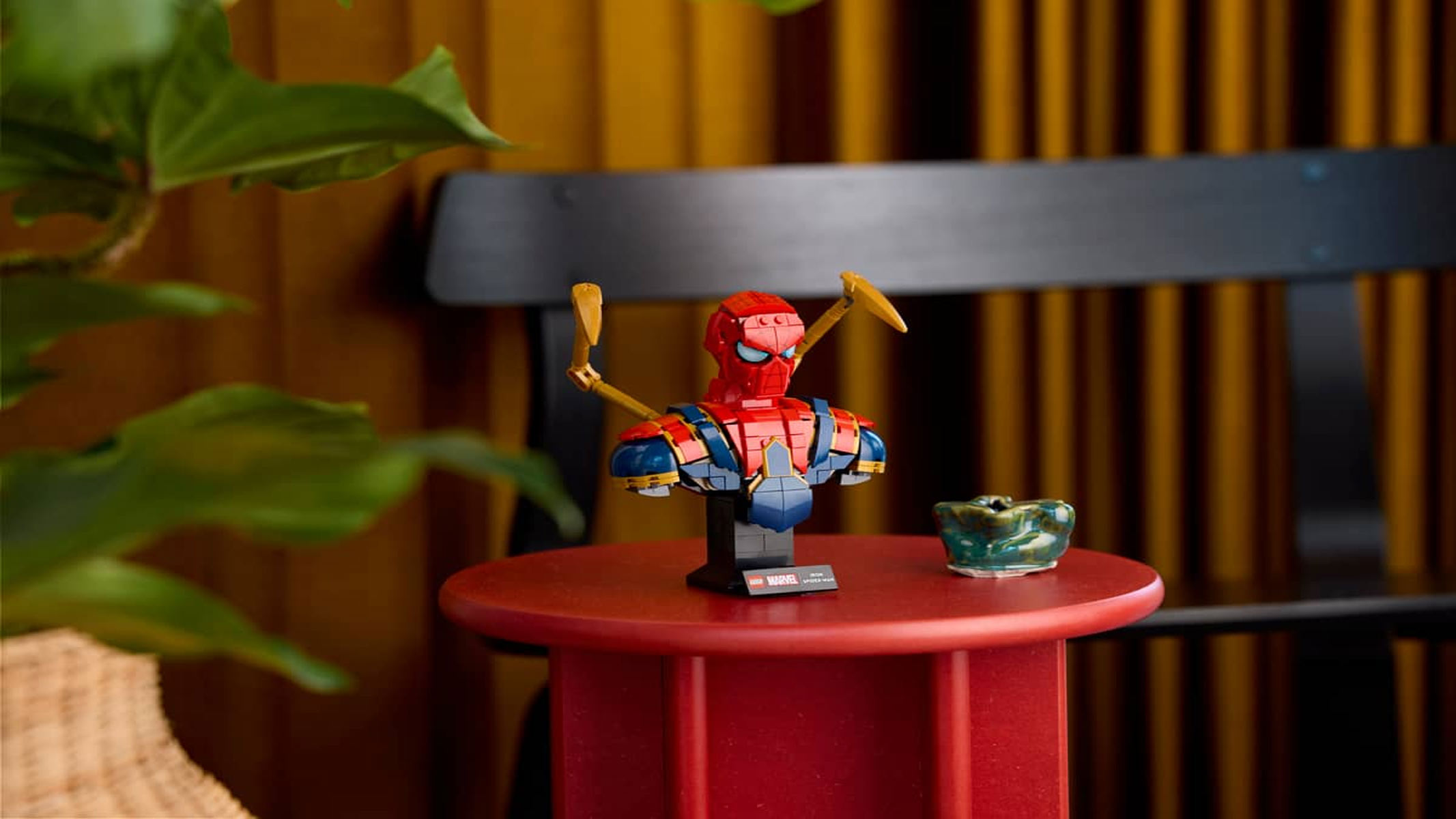 A red and blue bust of Spider-Man has gold arms coming out of the back, placed on a red table next to a plant on its left-hand side.