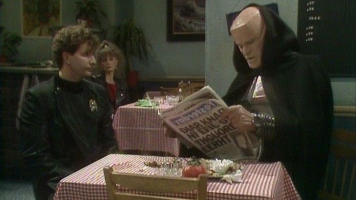 Still from the British sci-fi sitcom called Red Dwarf. Here we see Rimmer and Kryten in the 'Backwards' episodes. They are sitting in a cafe reading a backwards newspaper.