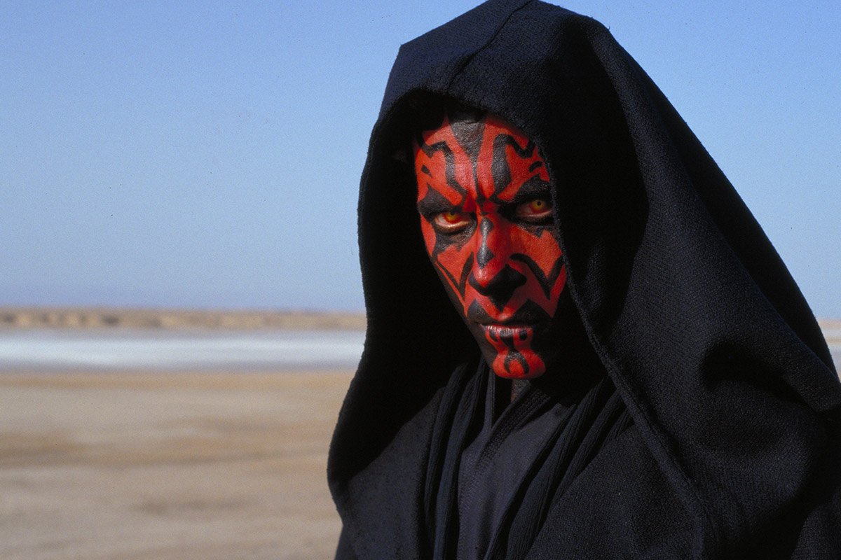 Darth Maul wearing a black hood, stood in a desert. This is a scene from Star Wars Episode 1: The Phantom Menace.