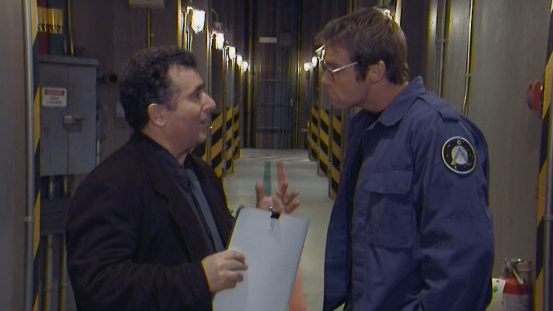 Screenshot from Stargate SG-1 showing two men having a conversation in a military corridor.
