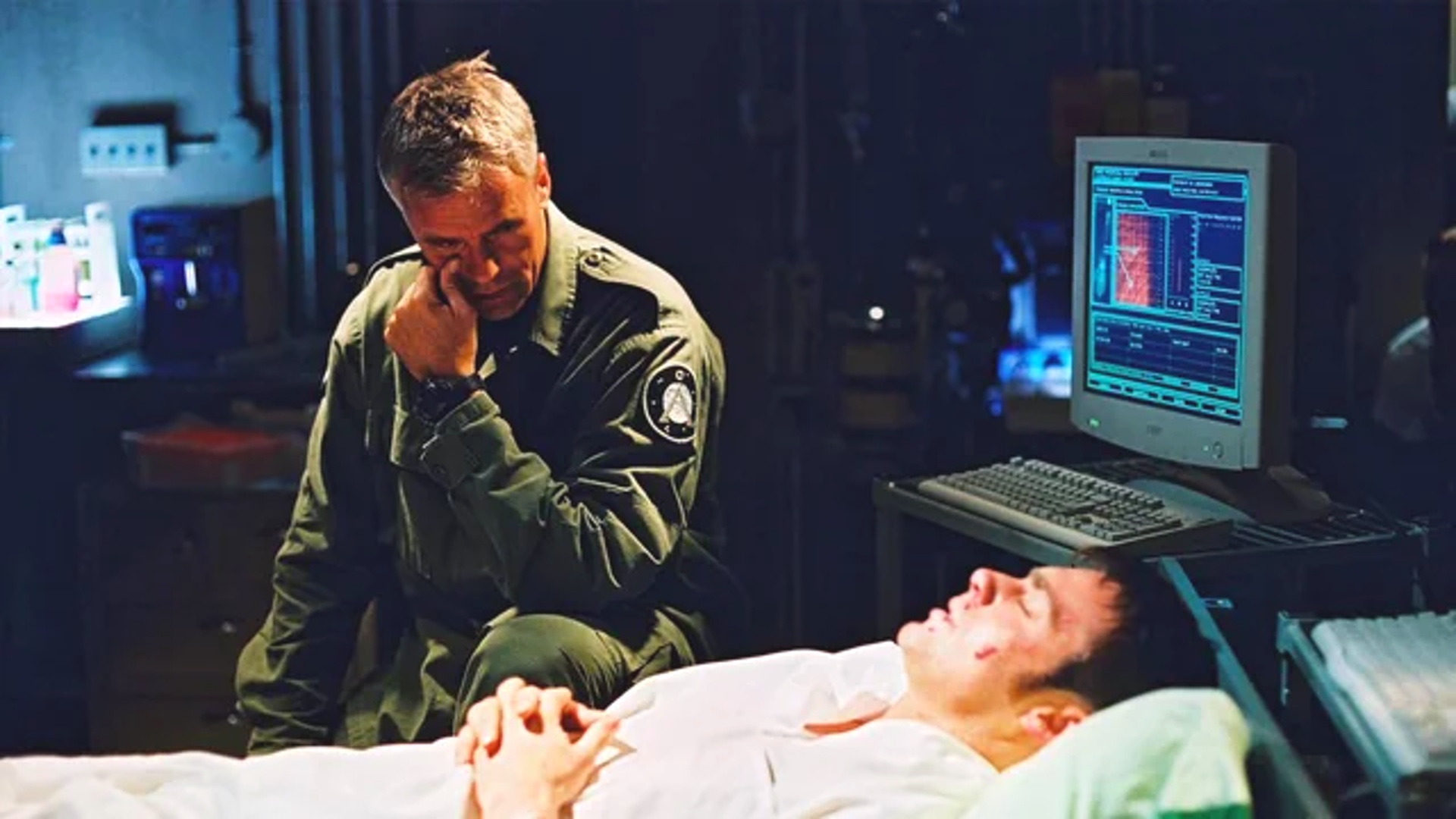 Screenshot from Stargate SG-1 showing a crying man sitting beside a dying man in a hospital bed.