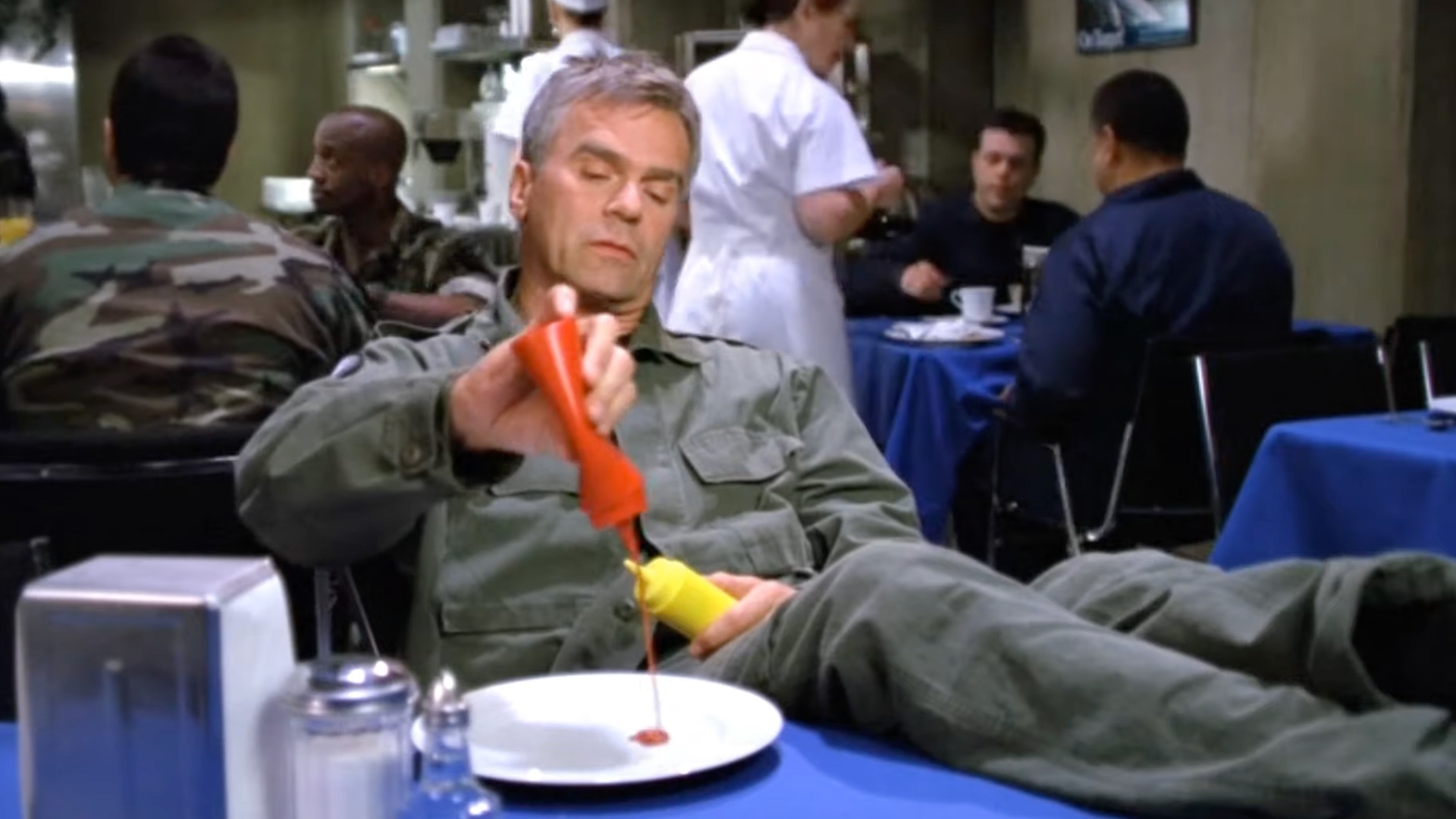 Screenshot from Stargate SG-1 showing a man squirting tomato ketchup onto a plate in a military base cafeteria.
